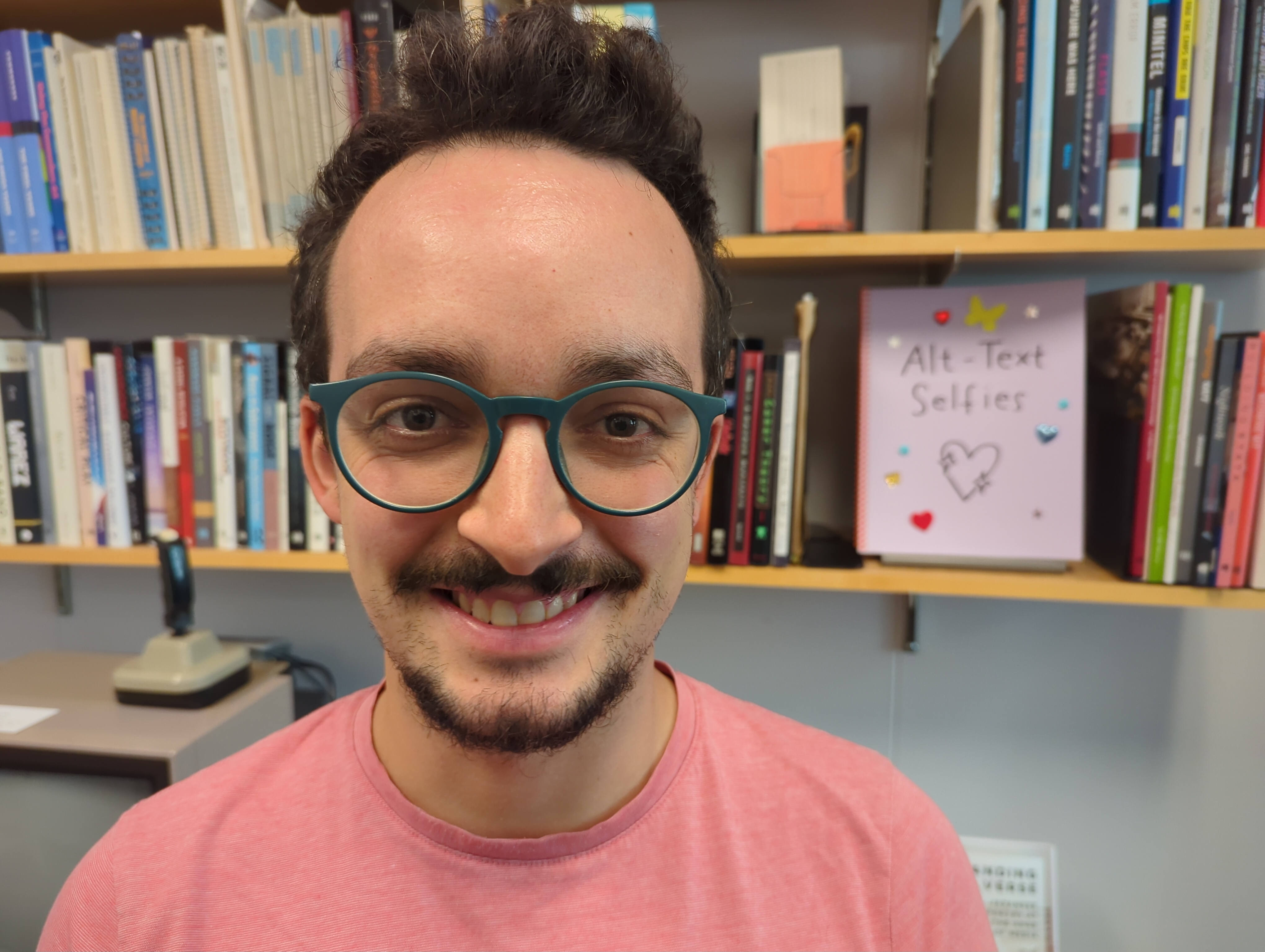 Louis is in roundish glasses and a peach shirt standing in front of a bookcase, and no, despite the booklet featured on the shelf, he didn’t write this ALT text himself!